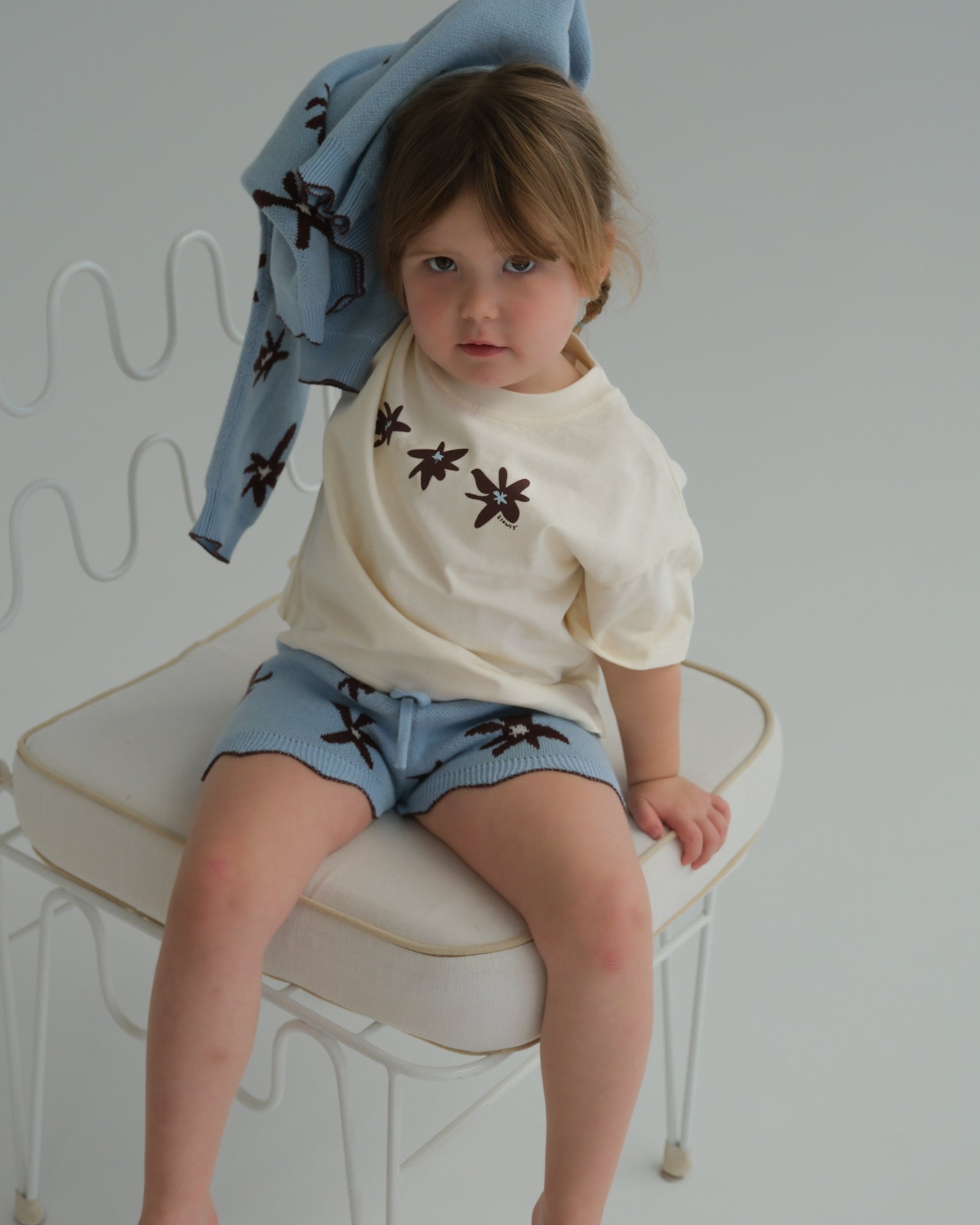 Child sitting on a chair wearing a white shirt with brown floral patterns and blue shorts.