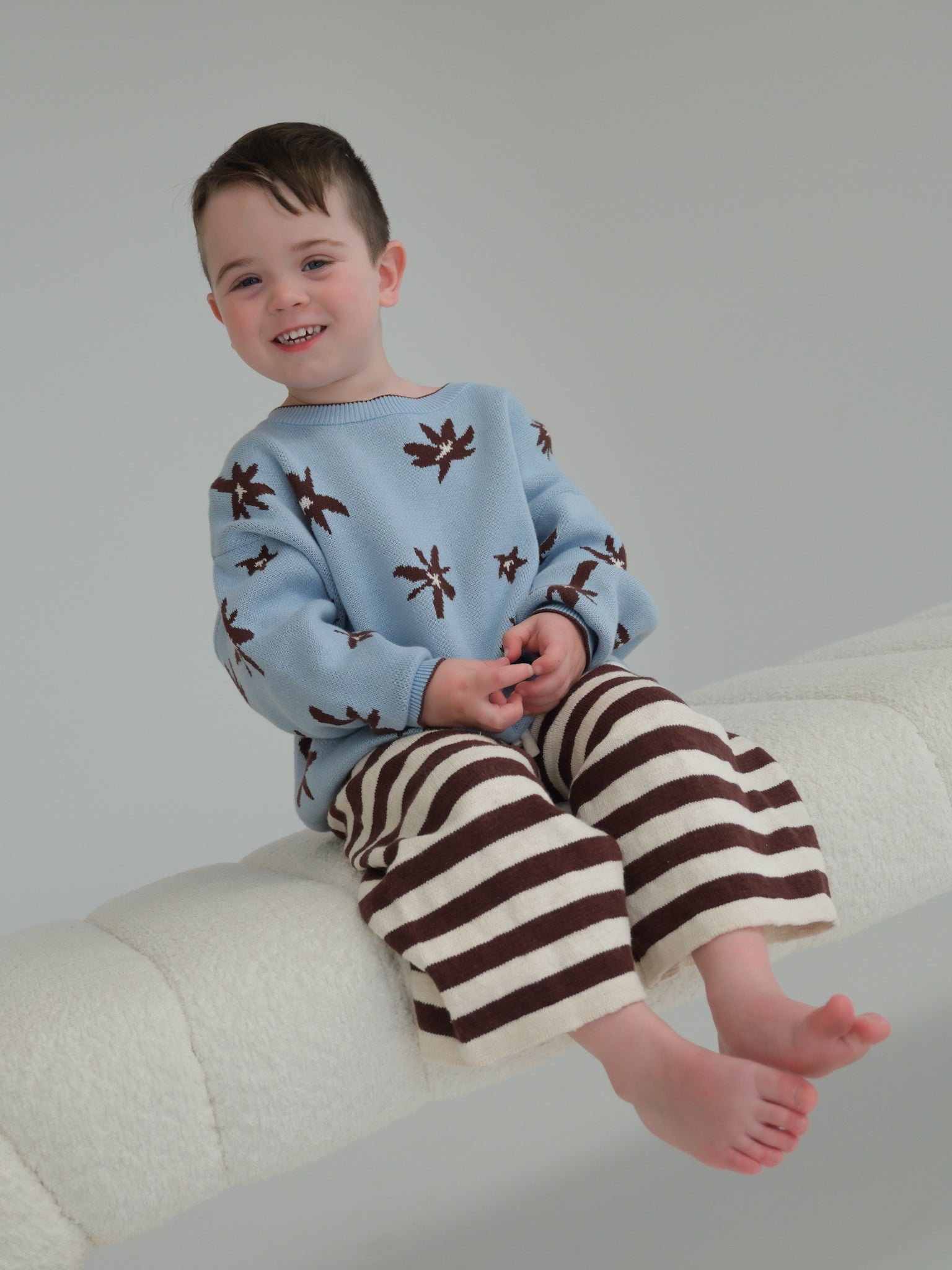 Child wearing a blue sweater with brown patterns and striped pants sitting on a white couch.